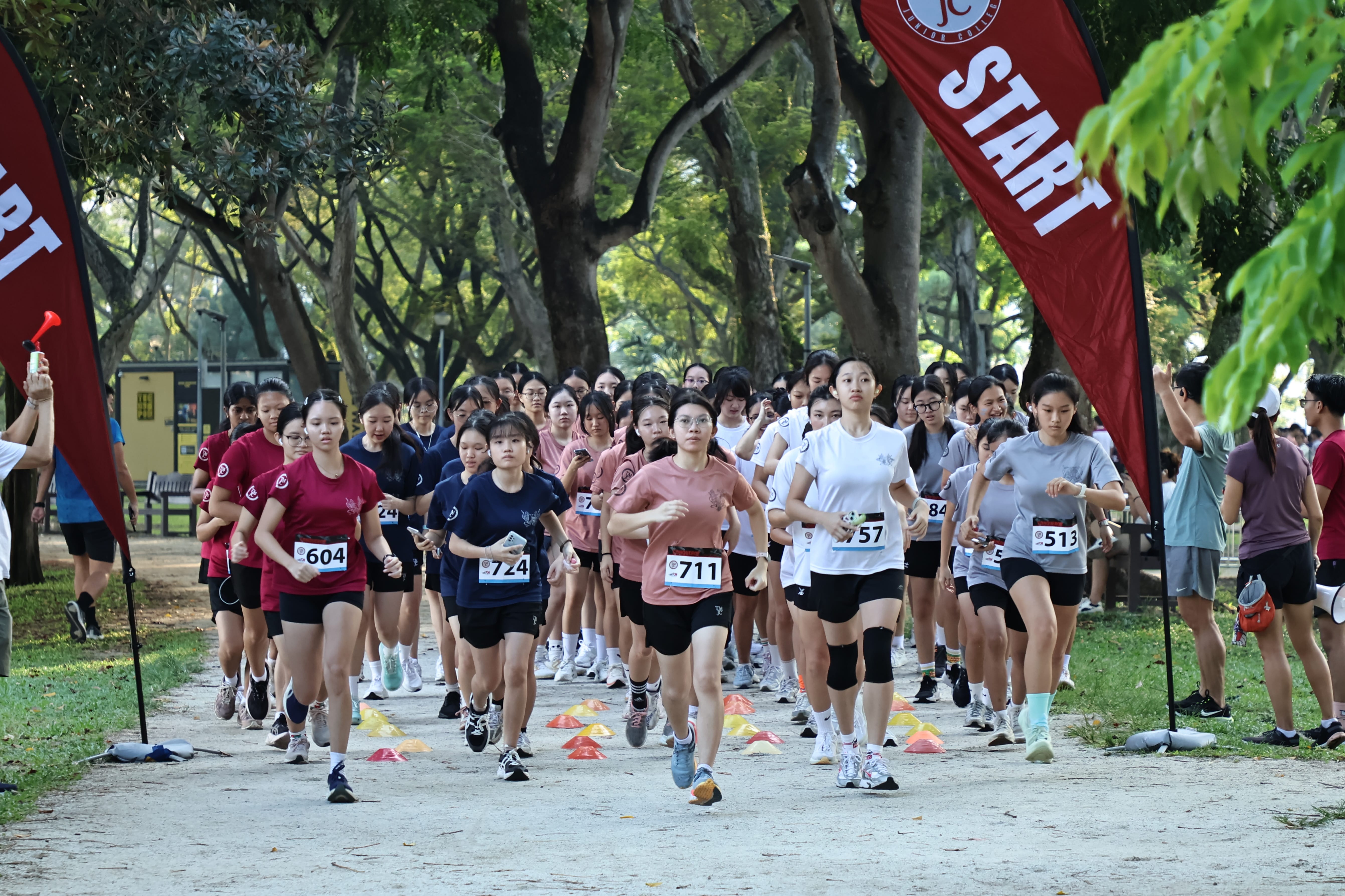 A large group of female students in various house-coloured T-shirts and race bibs surge forward from the start line of a 4km race along a tree-lined path.